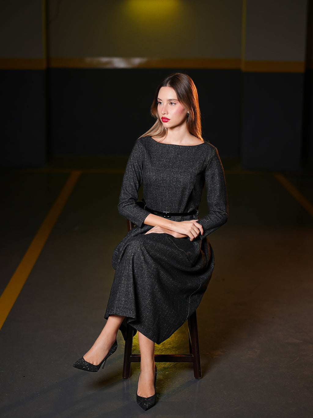 long-sleeve midi dress with a matching belt and black high heels, sitting on a wooden stool in a dimly lit indoor setting.