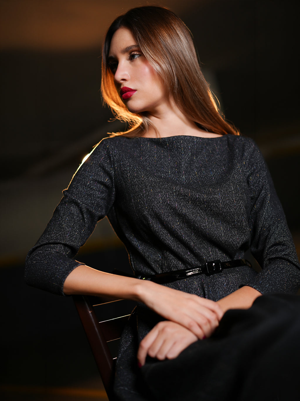 Dark charcoal gray textured three-quarter sleeve dress with a black belt, seated on a dark wooden chair against a dimly lit, moody background.