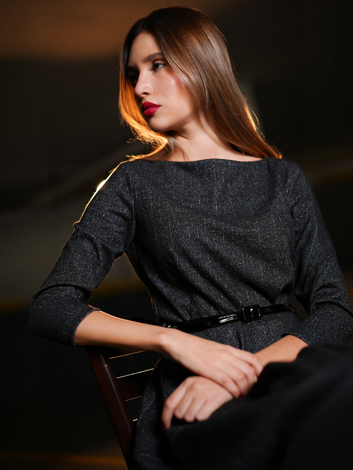 Dark charcoal gray textured three-quarter sleeve dress with a black belt, seated on a dark wooden chair against a dimly lit, moody background.