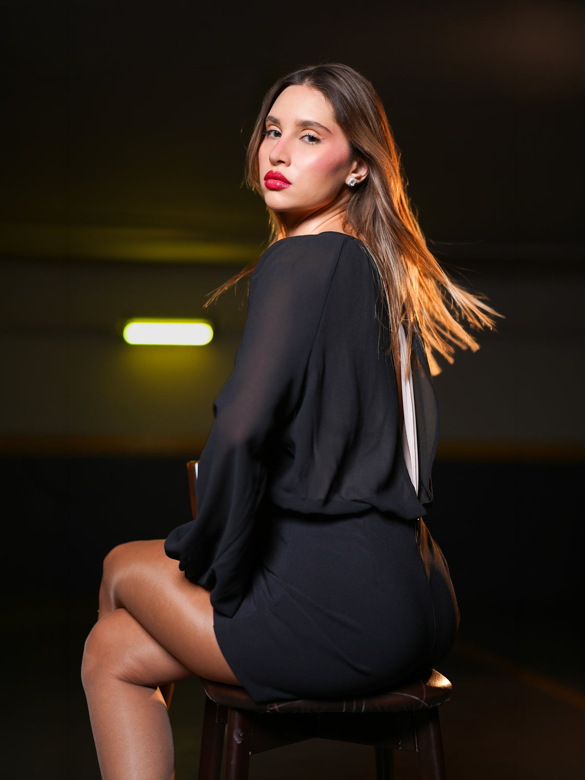 A woman sitting on a dark stool, wearing a sheer black long-sleeve overall.